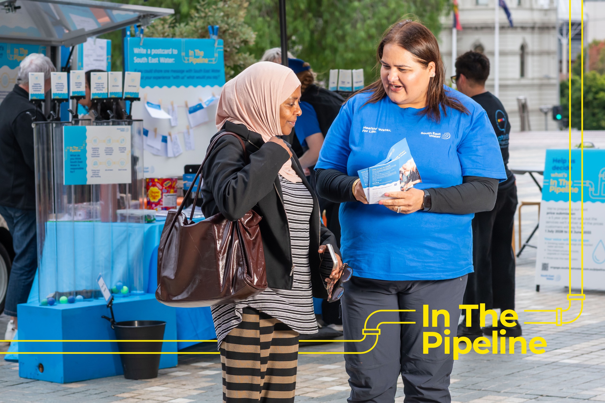 A staff member in a blue shirt speaks with a community member at an outdoor stall. They are holding information brochures, and event displays can be seen in the background.