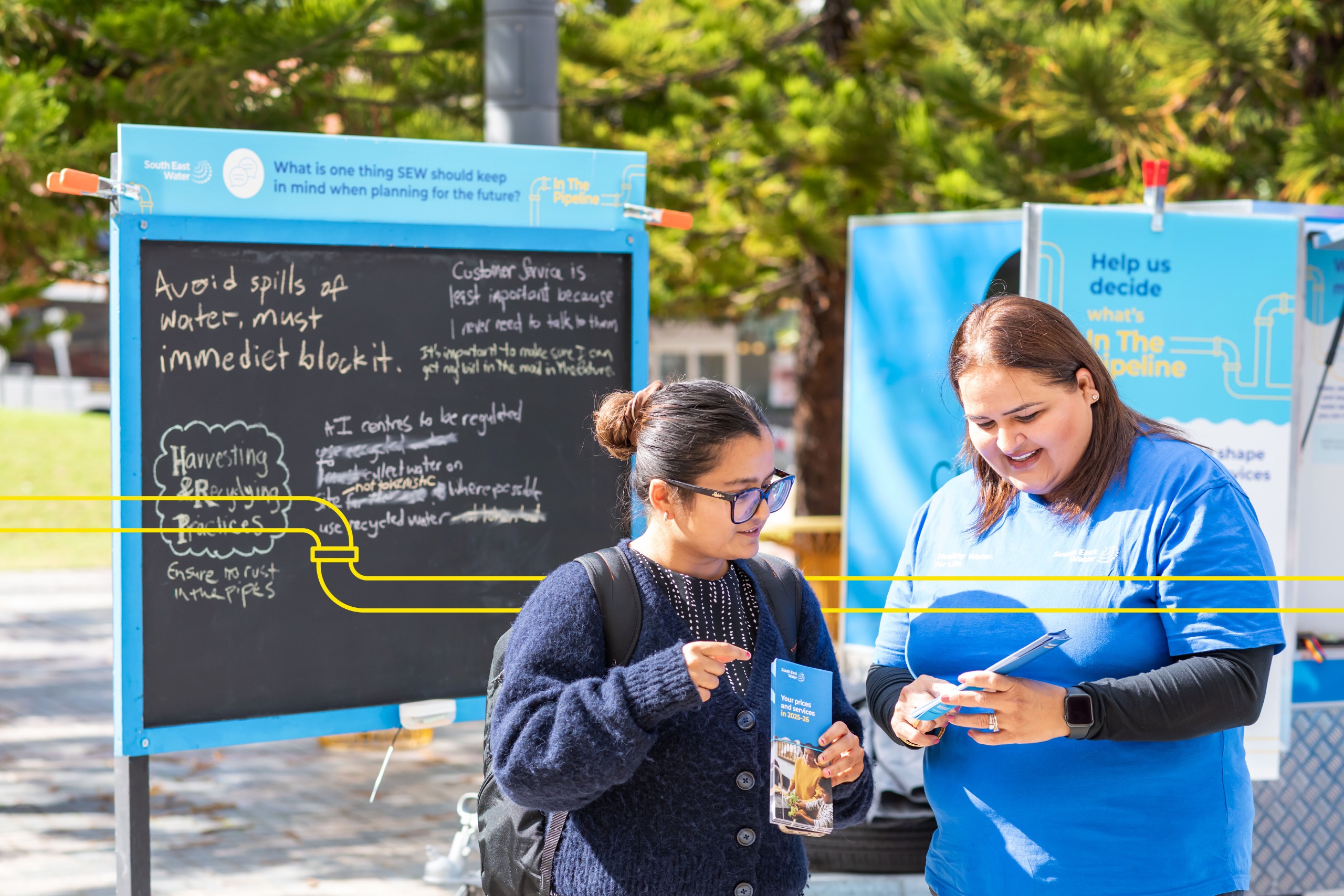 A staff member in a blue shirt talks with a community member outdoors. Behind them is a chalkboard with ideas about water services written on it.