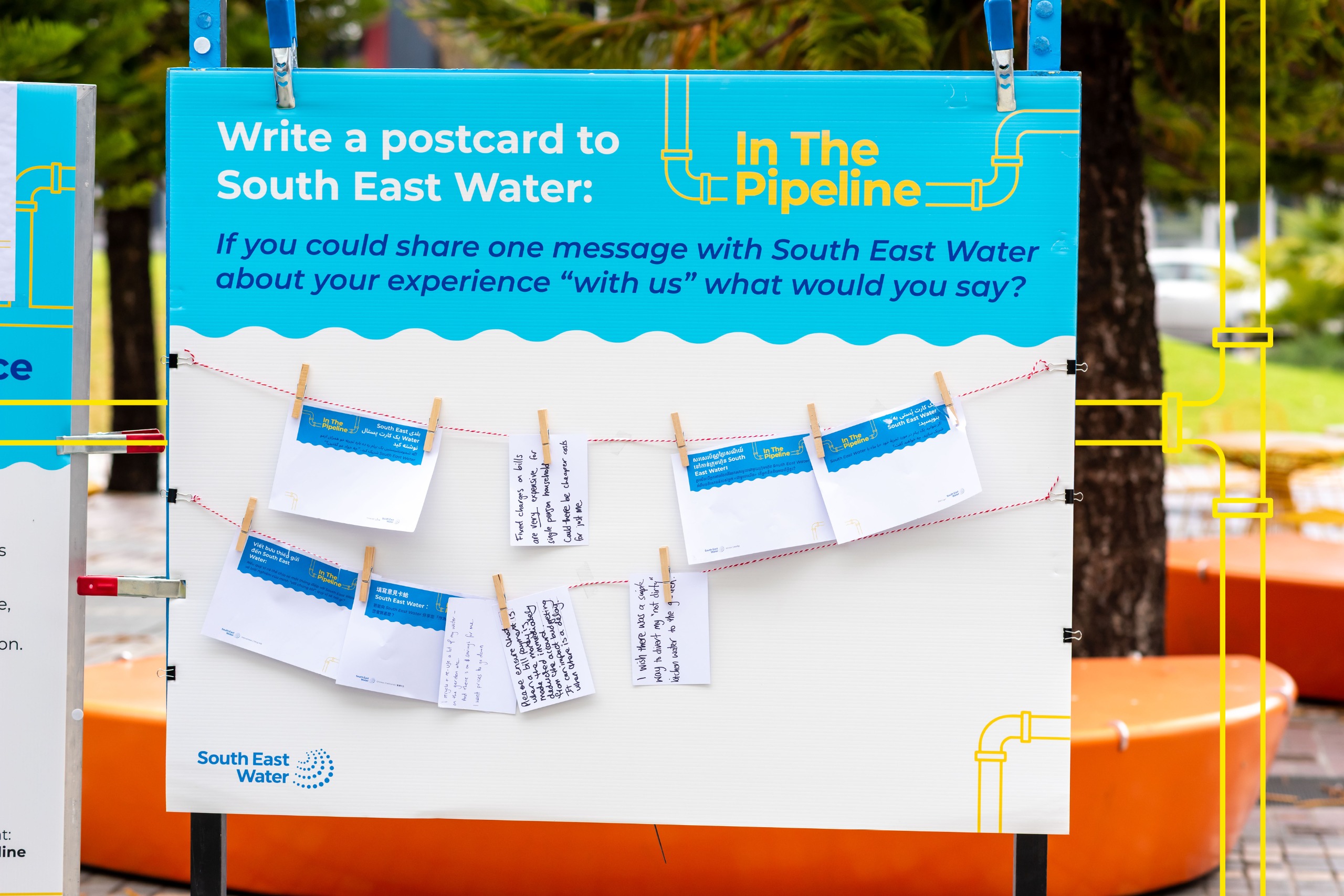 A display board says “Write a postcard to South East Water.” Below the heading, several small postcards are clipped to string. People have written short messages on the postcards about their experience with South East Water.
